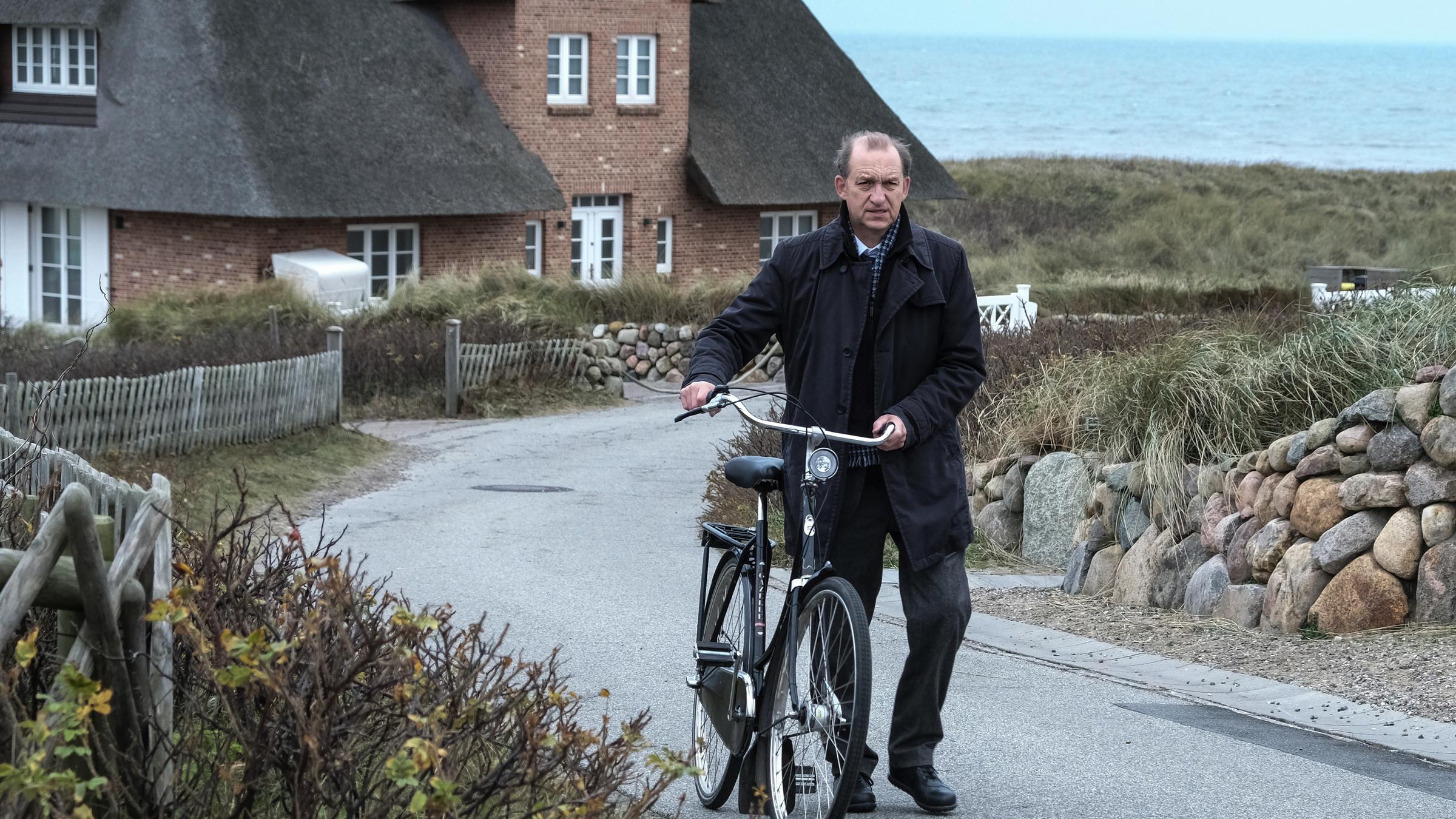 „Nord Nord Mord – Sievers und die Frau im Zug“: Carl Sievers (Peter Heinrich Brix) schiebt das Fahrrad die Straße hinauf, im Hintergrund sind ein reetgedecktes Backsteinhaus mit weißen Fenster, Gartenmauern und -zäune sowie das Meer zu sehen.
