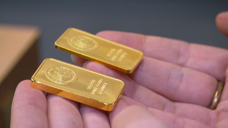"Falsche Versprechen? So lassen wir uns manipulieren": Nahaufnahme von zwei kleinen Goldbarren, die auf den Fingern einer Person liegen.