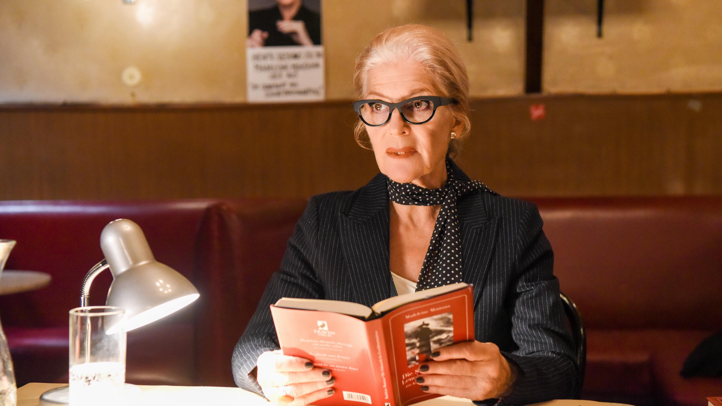 "Die Muse des Mörders": Madeleine Montana (Christiane Hörbiger) sitzt mit einem aufgeschlagenen Buch an einem Tisch. Neben ihr stehen ein Glas Wasser und eine Leselampe.