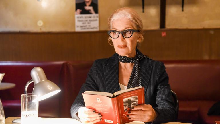 "Die Muse des Mörders": Madeleine Montana (Christiane Hörbiger) sitzt mit einem aufgeschlagenen Buch an einem Tisch. Neben ihr stehen ein Glas Wasser und eine Leselampe.