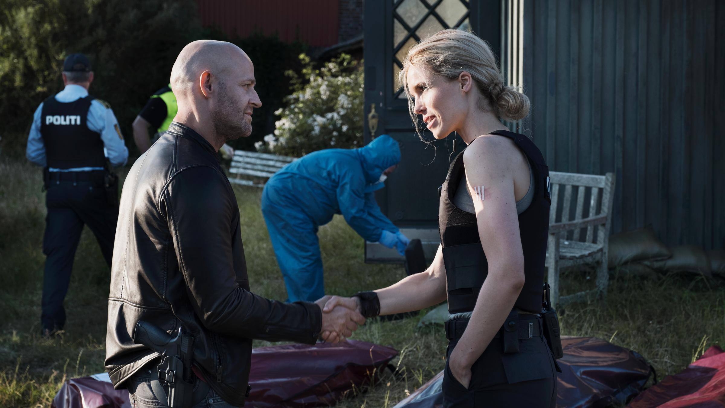 "The Team II": Gregor Weiss (Jürgen Vogel) und Nelly Winther (Marie Bach Hansen) geben sich die Hand. Im Hintergrund sieht man einen Tatort und Mitarbeiter der Polizei.