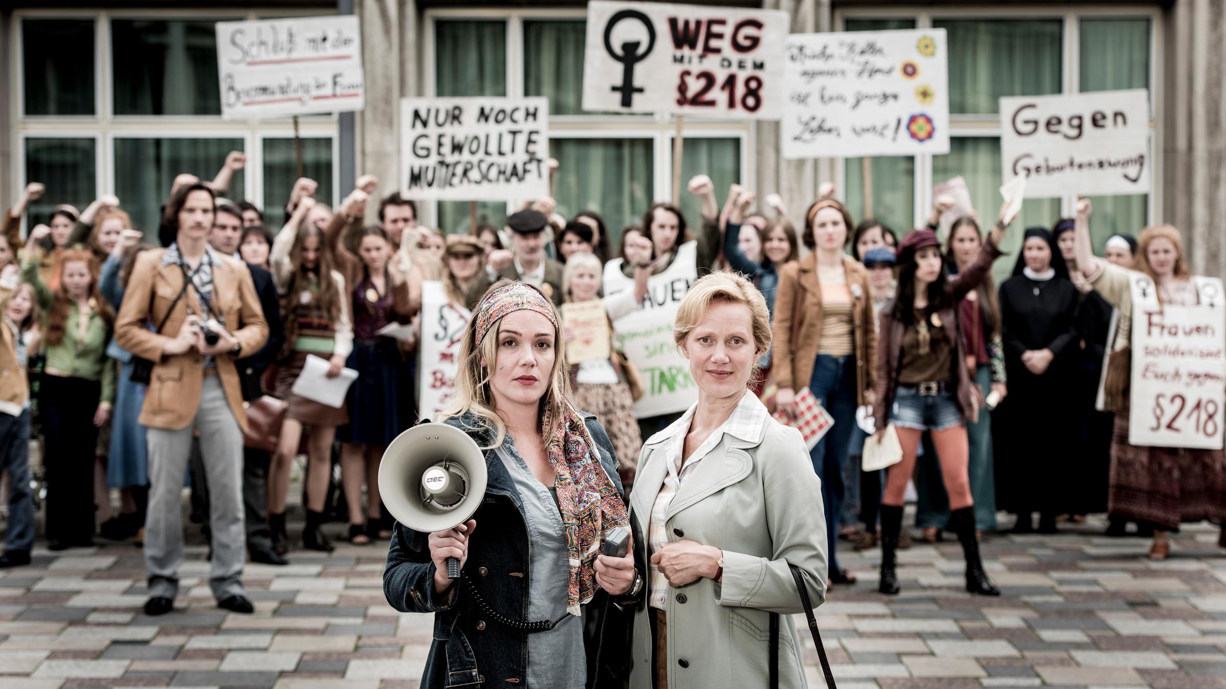 "Aufbruch in die Freiheit": Charlotte (Alwara Höfels) und Erika (Anna Schudt) stehen mit einem Megaphon vor einer demonstrierenden Menschenmenge. Die Frauen und Männer halten Plakate hoch und heben protestierend ihre Fäuste.