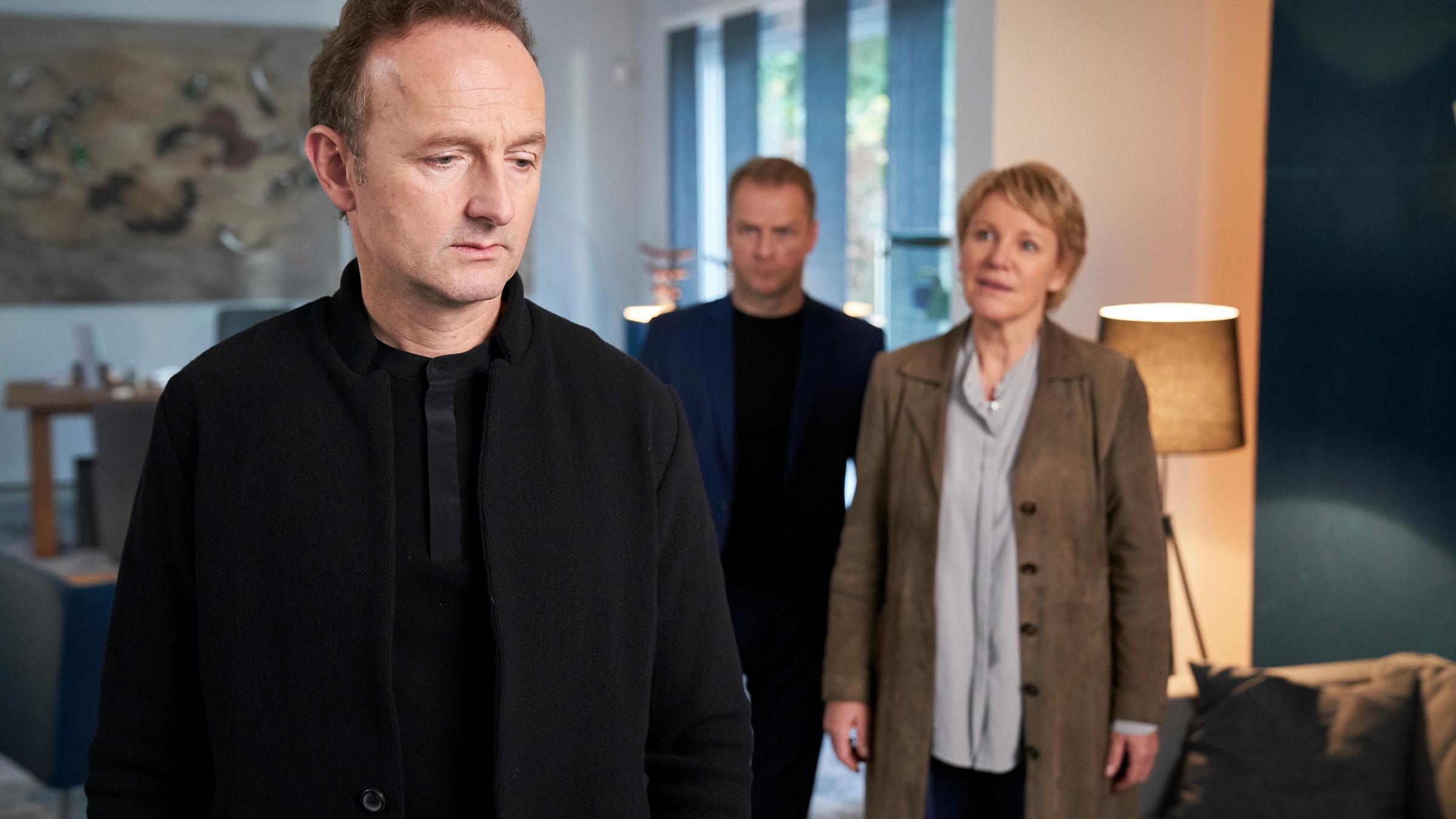 "Marie Brand und das Verhängnis der Liebe": Jörg Korfmann (Peter Jordan) steht im Wohnzimmer seines Hauses, hinter ihm stehen Jürgen Simmel (Hinnerk Schönemann) und Marie Brand (Mariele Millowitsch). Während Marie Korfmann etwas sagt, hören sie ihm aufmerksam zu.