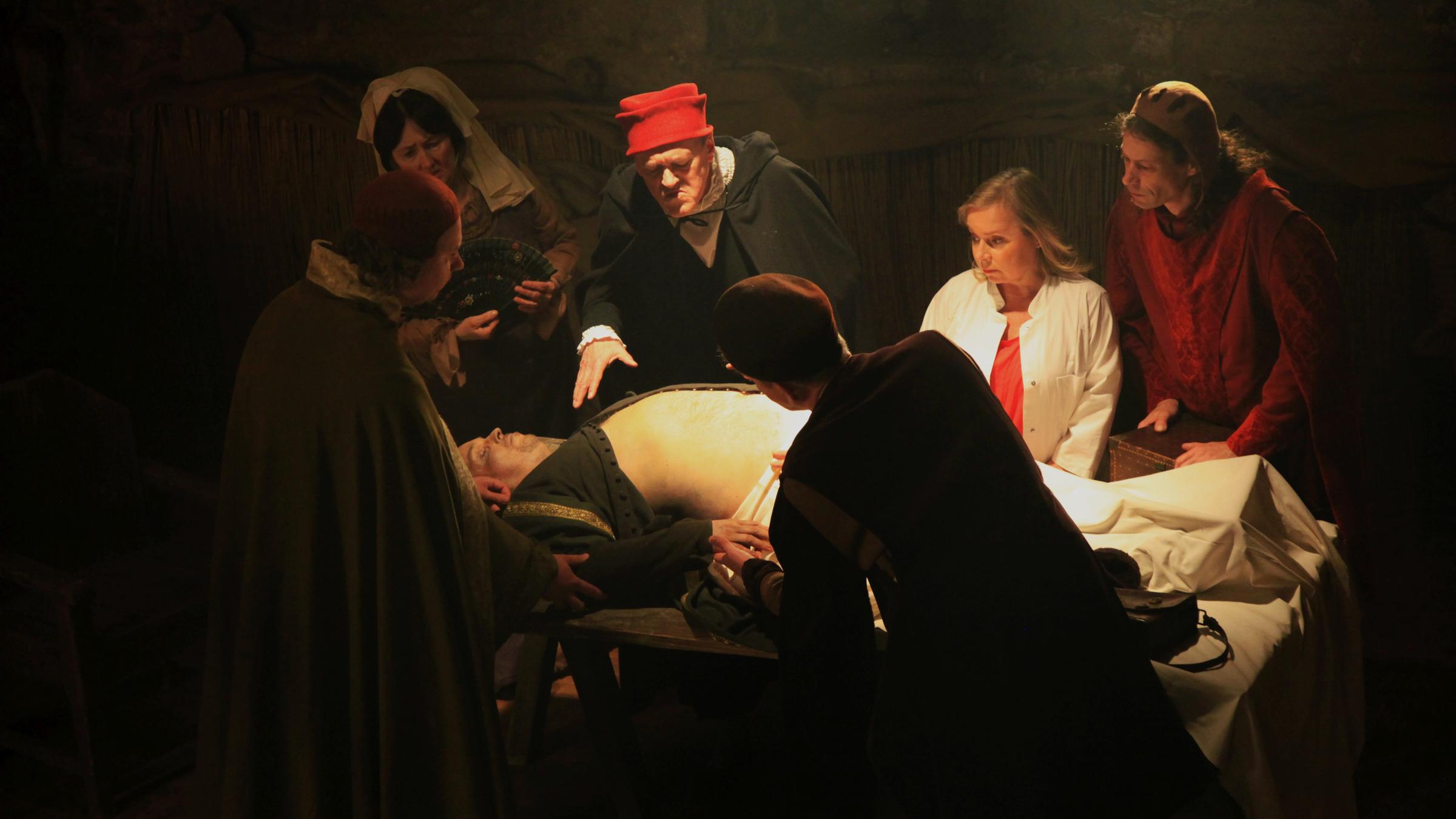 "Verräterische Spuren - Die Geschichte der Forensik: Was Opfer preisgeben": Dunkler Hintergrund, im Vordergrund ein hell beleuchteter Tisch: Mehrere Darsteller, unter ihnen ChrisTine Urspruch, stehen um den Tisch  und betrachten einen darauf liegenden Mann mit entblößtem Bauch.