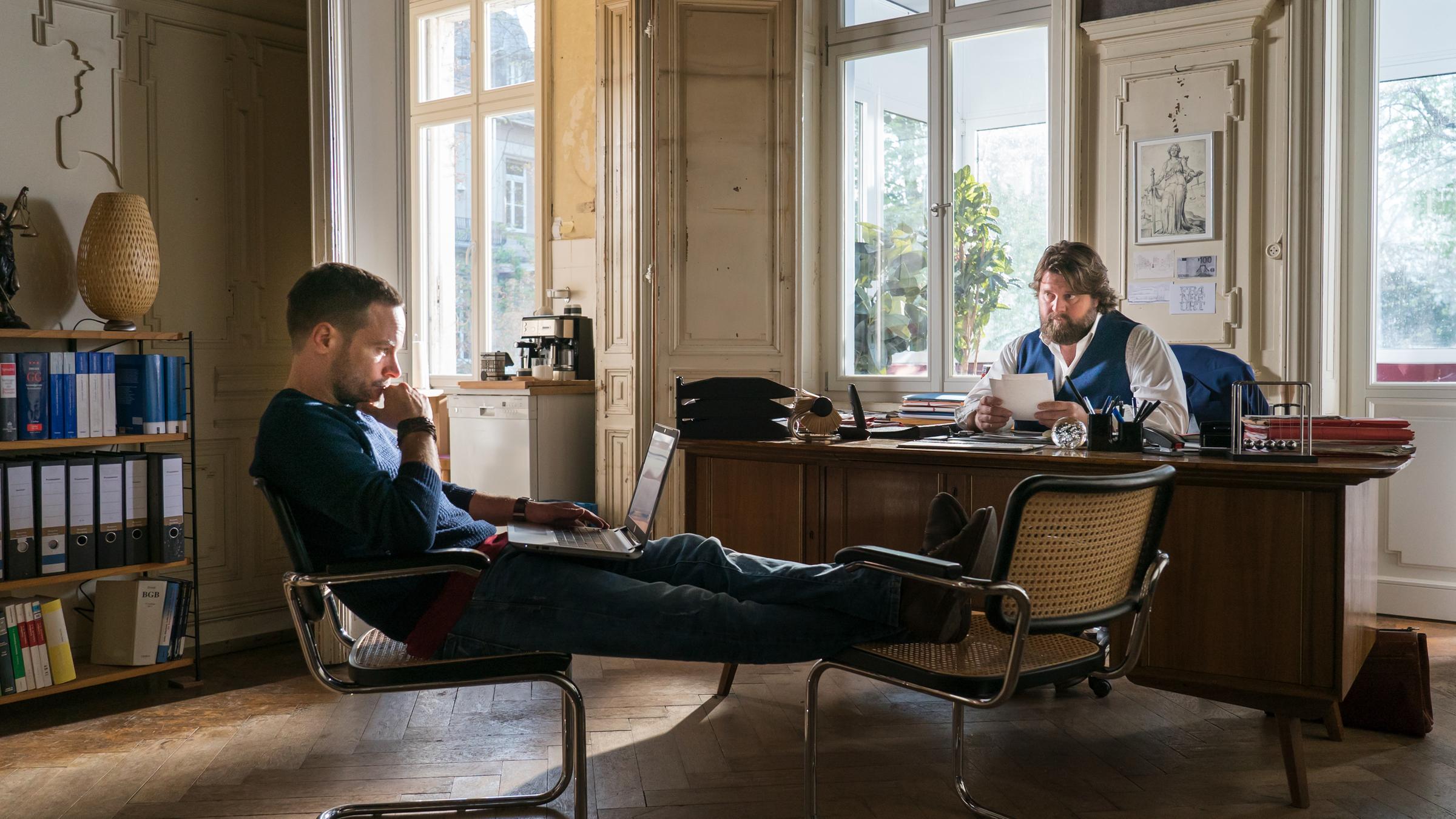 „Ein Fall für zwei – Familienbande“:  Leo Oswald (Wanja Mues) sitzt auf einem Stuhl, die Beine auf einem weiteren Stuhl hochgelegt, vor Bennis (Antoine Monot, Jr.) Schreibtisch. Er hat einen Laptop auf dem Schoß und schaut nachdenklich auf dessen Bildschirm. Benni sitzt hinter seinem Schreibtisch und blickt erwartungsvoll zu Leo.