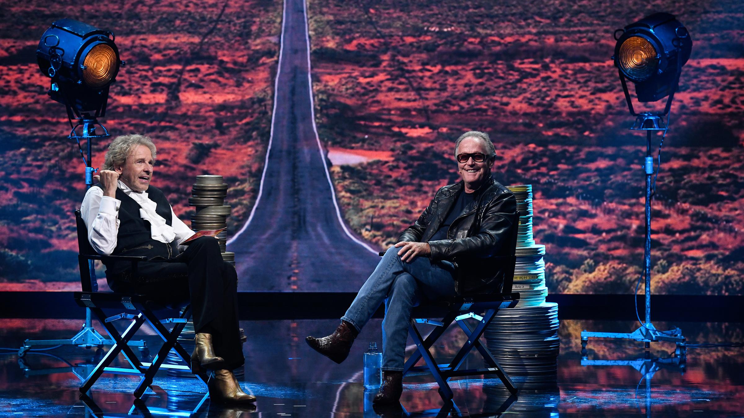 "Gottschalks große 68er-Show": Thomas Gottschalk sitzt mit der Hollywood-Legende Peter Fonda vor einer Leinwand auf der eine scheinbar endlose Asphaltpiste abgebildet ist.