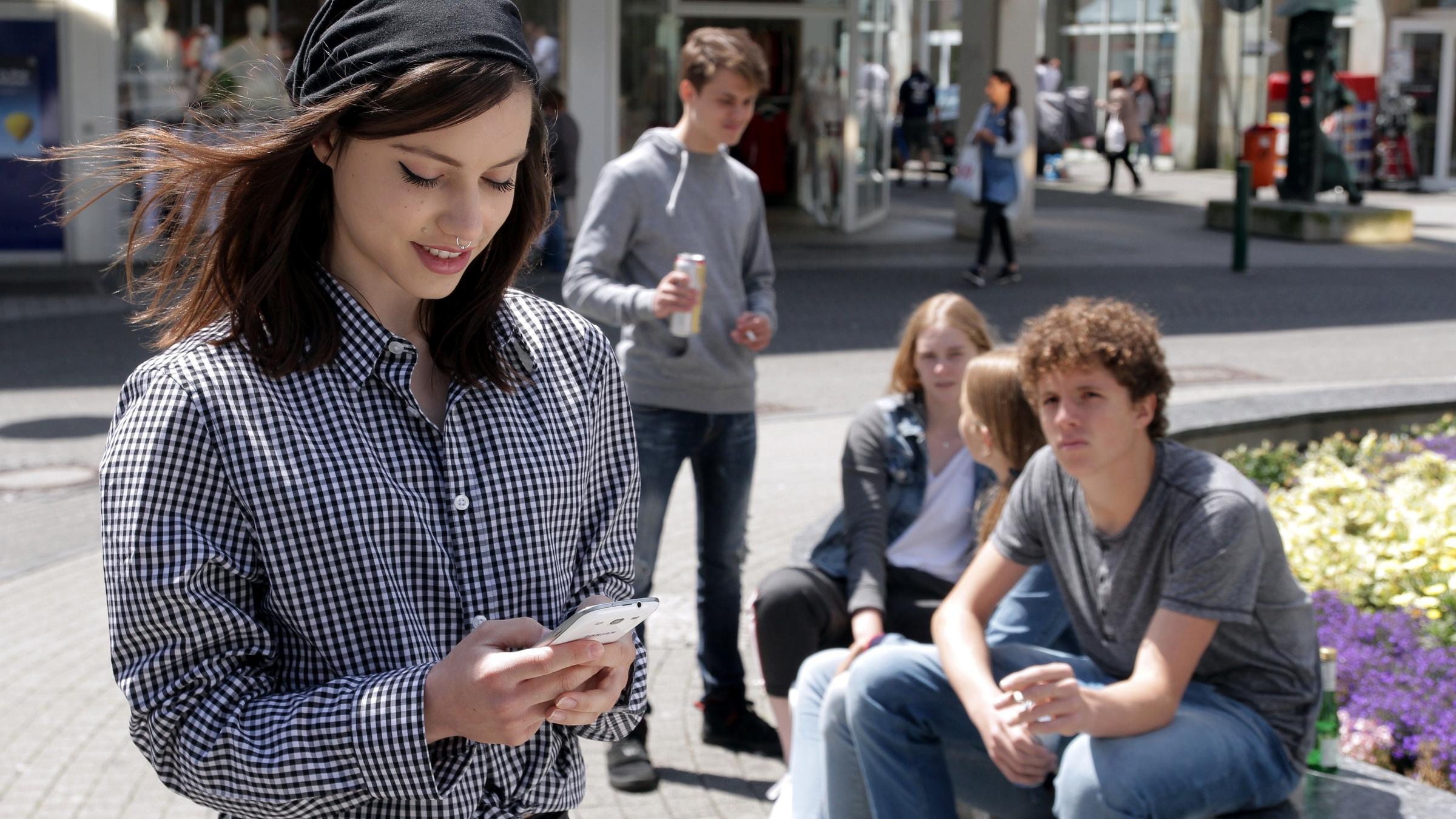 "Aktenzeichen XY... ungelöst - Spezial": Ein junges Mädchen läuft durch eine Fußgängerzone und schaut dabei auf ihr Handy, sie lächelt. Dahinter sieht man noch andere Jugendliche die sich unterhalten. Einer von ihnen hält eine Zigarette in der Hand und schaut sich nach ihr um.