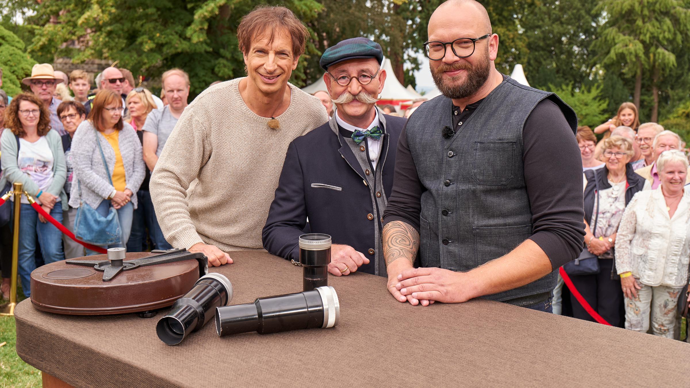 "Bares für Rares - Deutschlands größte Trödelshow": Ingolf Lück, Horst Lichter und Sven Deutschmanek stehen dicht nebeneinander hinter einem Tisch lächeln in die Kamera.