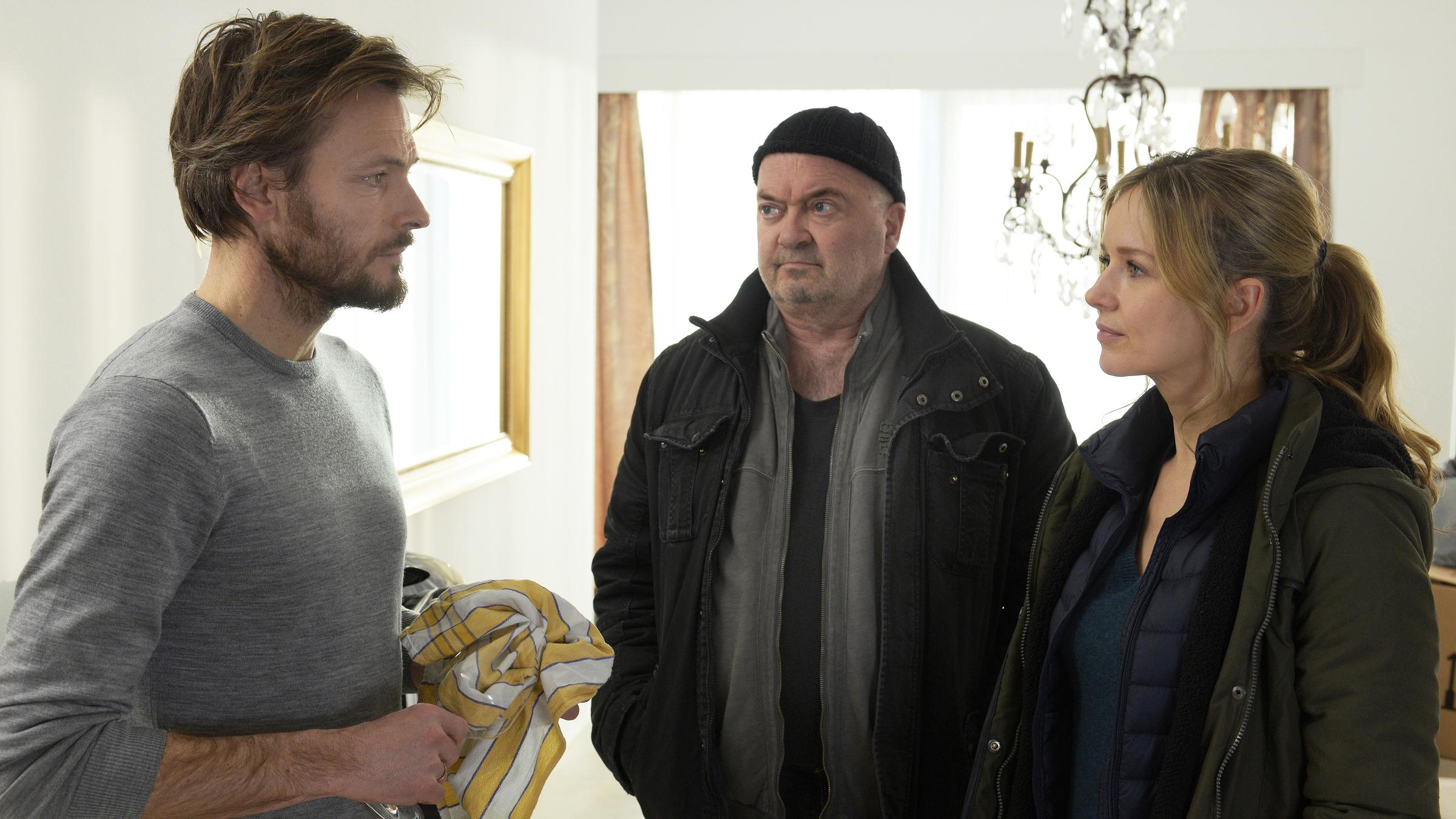 "Ein starkes Team – Tödlicher Seitensprung": Michael Sanders (Andreas Pietschmann), der Ehemann des Opfers), redet mit den Ermittlern Linett (Stefanie Stappenbeck) und Otto (Florian Martens). Die drei befinden sich in einem Wohnzimmer. Sanders trocknet gerade ein Glas ab und schaut Linett intensiv an. Otto beobachtet Sanders.