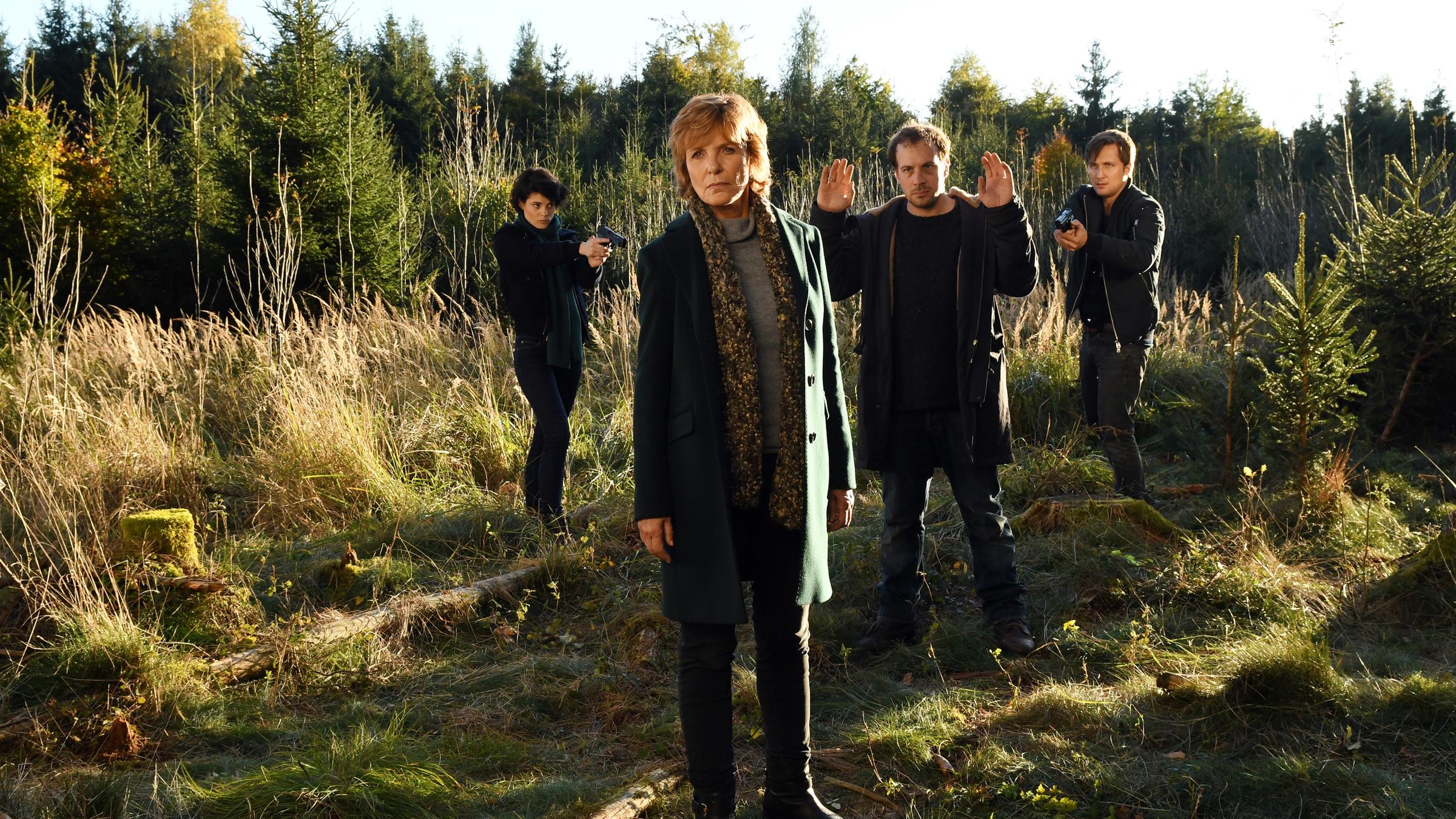 “Kommissarin Lucas – Das Urteil“: Am Waldrand. Tom Brauer (Lasse Myhr) und Judith Marlow (Jördis Richter) zielen mit ihren Waffen auf den vor ihnen stehenden Bernd Stach (Florian Teichtmeister) der beide Hände hochhält. Bernd Stach blickt in Richtung Ellen Lucas (Ulrike Kriener), die mit dem Rücken zu den anderen vor ihnen steht und nach vorne blickt.