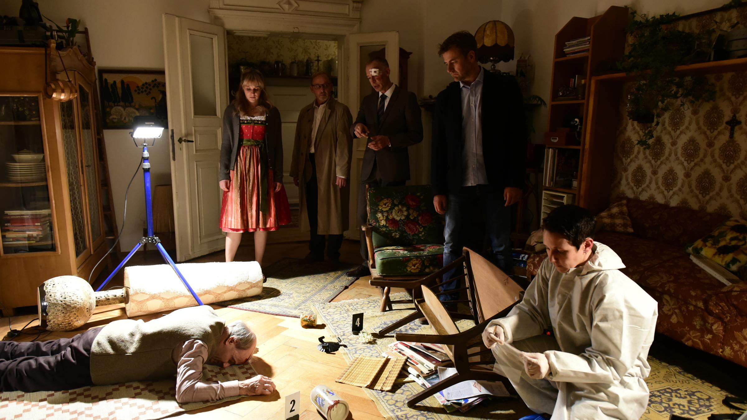  „München Mord – Die ganze Stadt ein Depp“: Angelika (Bernadette Heerwagen), Schaller (Alexander Held), Zangel (Christoph Süß) und Harald (Marcus Mittermeier) stehen in Josef Kleints (Nikolaus Paryla) Wohnzimmer und sehen sich den Tatort an. Das Mordopfer liegt am Boden.