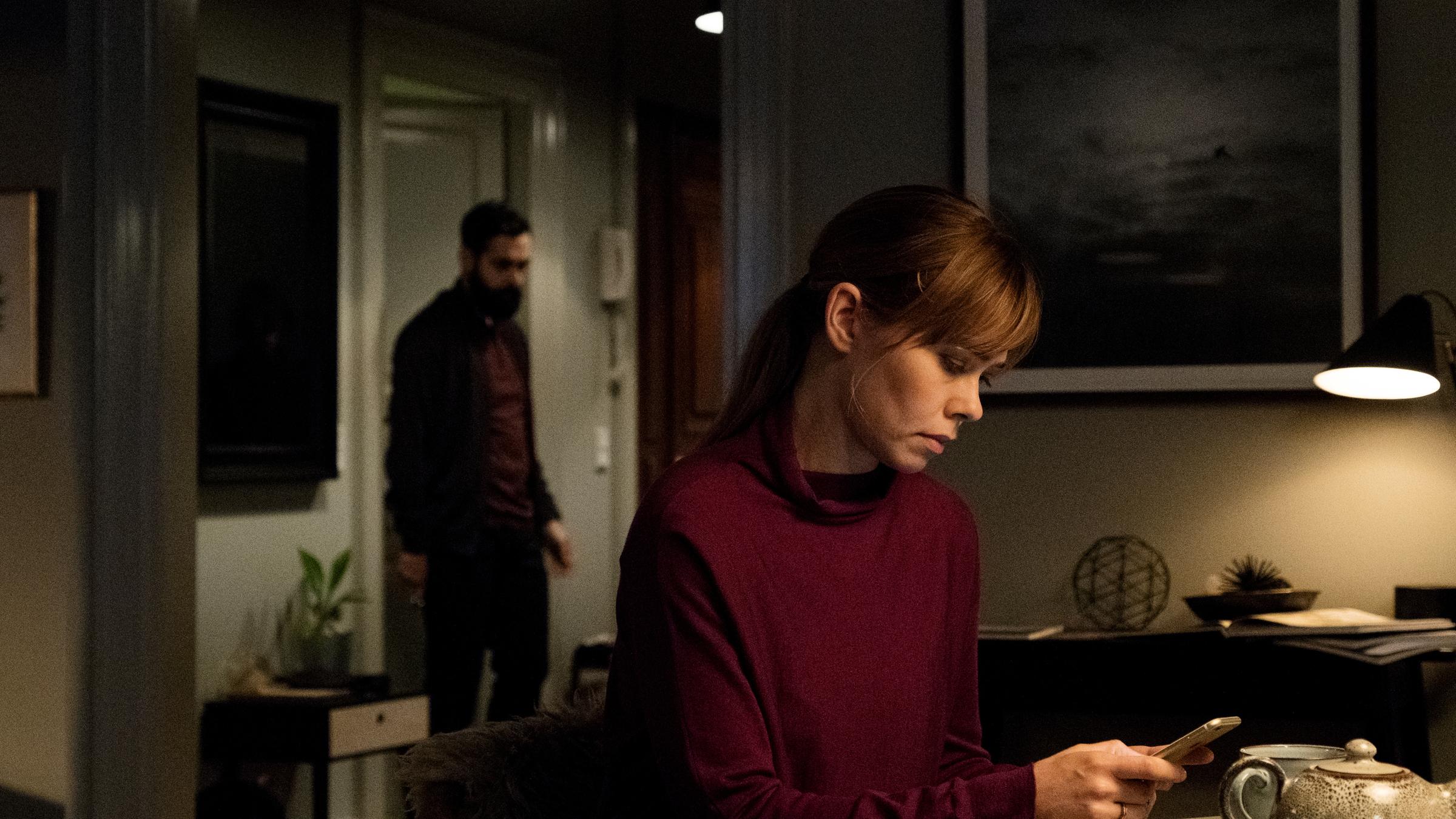 "Greyzone - No Way out: Die Vereinbarung": Victoria (Birgitte Hjort Sørensen) sitzt an einem Esstisch und blickt konzentriert auf ihr Handy. Im Flur ist leicht unscharf Iyad (Ardalan Esmali) zu erkennen.