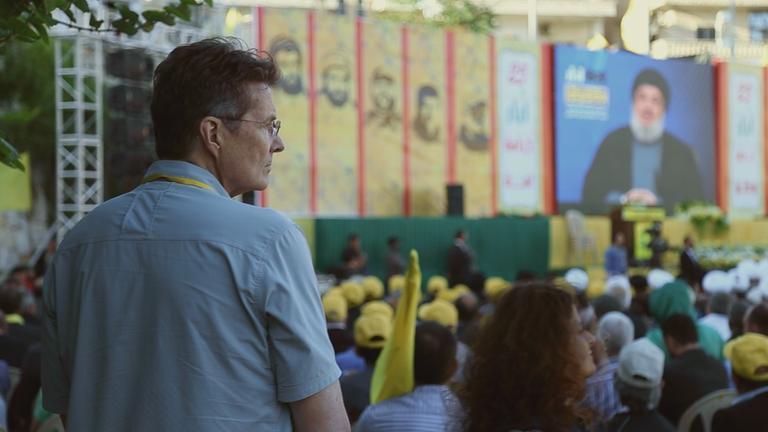 "Öl, Macht und Religion - Saudi-Arabien und der Iran - Alte Rivalen": Reporter Martin Smith steht hinter einer großen Menschengruppe, die in die Richtung einer Videoleinwand schaut, auf der ein Redner mit Bart und Turban zu sehen ist.