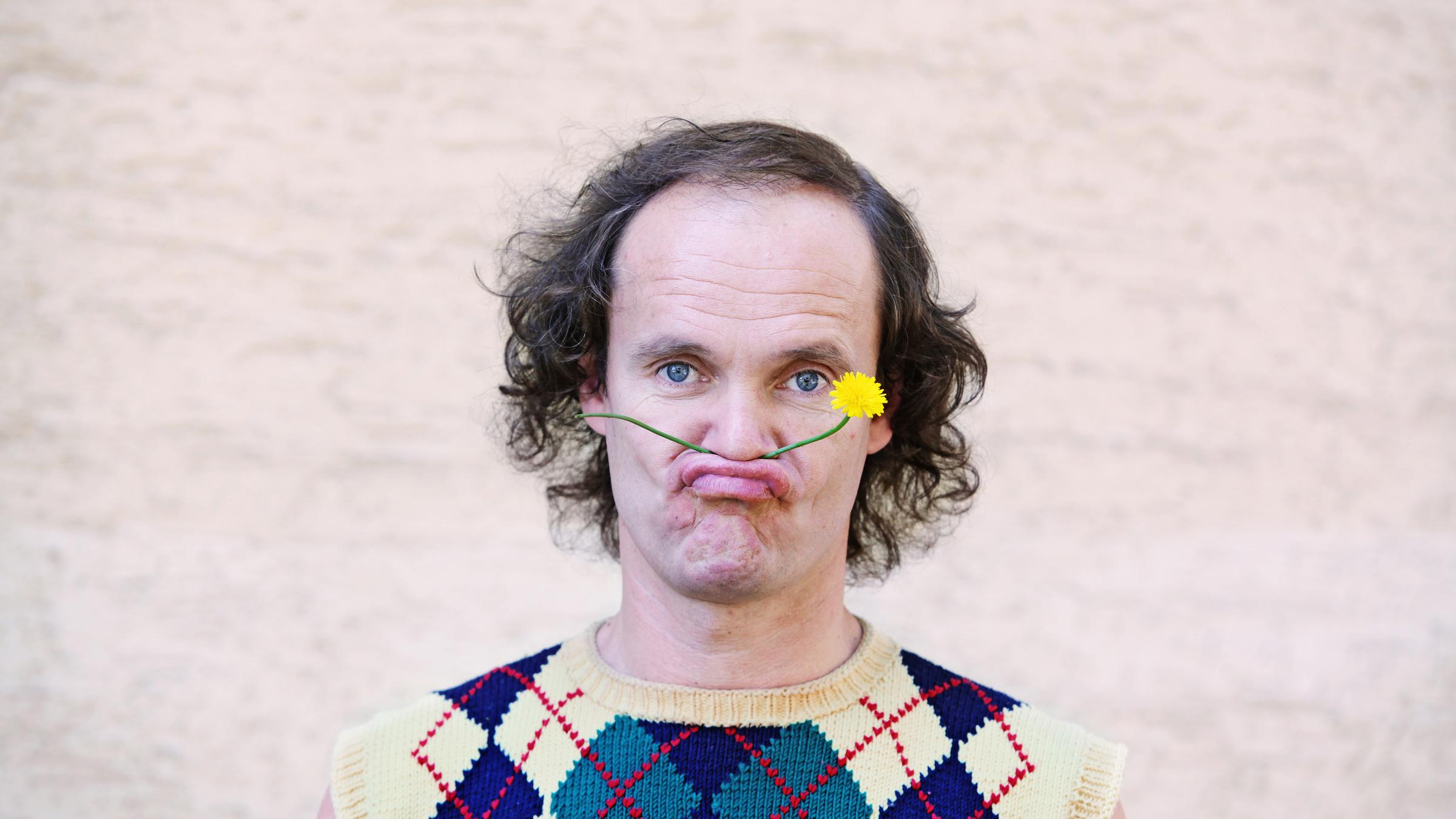 "Olaf Schubert: Sexy forever": Portrait von Olaf Schubert. Er hat sich eine gelbe Löwenzahnblume zwischen Oberlippe und Nase geklemmt und schaut in die Kamera.