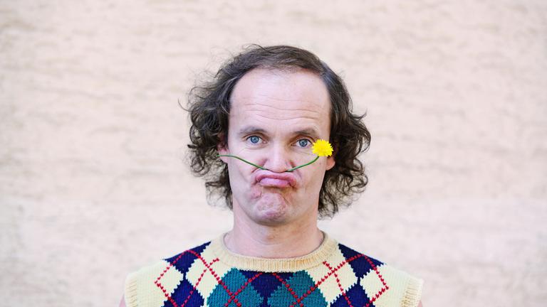 "Olaf Schubert: Sexy forever": Portrait von Olaf Schubert. Er hat sich eine gelbe Löwenzahnblume zwischen Oberlippe und Nase geklemmt und schaut in die Kamera.