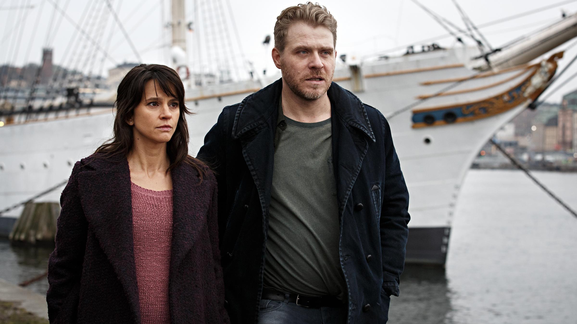 "Anne Holt: Der Mörder in uns": Psychologin Johanne Vik (Melinda Kinnaman) und Kommissar Ingvar Nyman (Henrik Norlén) laufen nebeneinander auf einem Kai im Hafen. Im Hintergrund ist ein Segelschiff zu sehen.