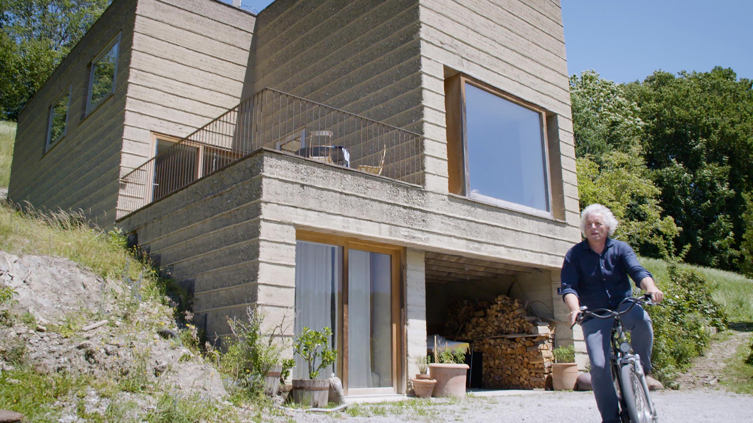 ""plan b: Das Haus von Morgen - Wie Bauen revolutioniert wird": Lehmbauer Martin Rauch sitzt vor seinem modernen Haus aus Erde auf dem Fahrrad.