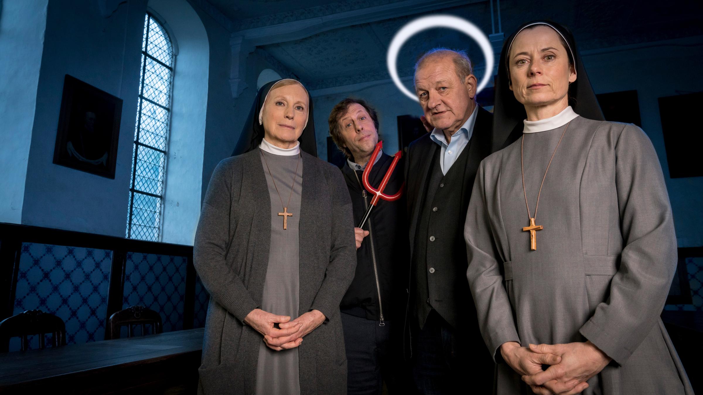 "Wilsberg: Gottes Werk und Satans Kohle": Ekki Talkötter (Oliver Korittke) hält einen Satans-Dreizack in der Hand, und Georg Wilsberg (Leonard Lansink) hat einen Heiligenschein über dem Kopf. Schwester Helena (Maren Kroymann) und Schwester Christa (Inka Friedrich) stehen in Habit und mit gefalteten Händen neben den beiden in der Kirche. Alle schauen in die Kamera.