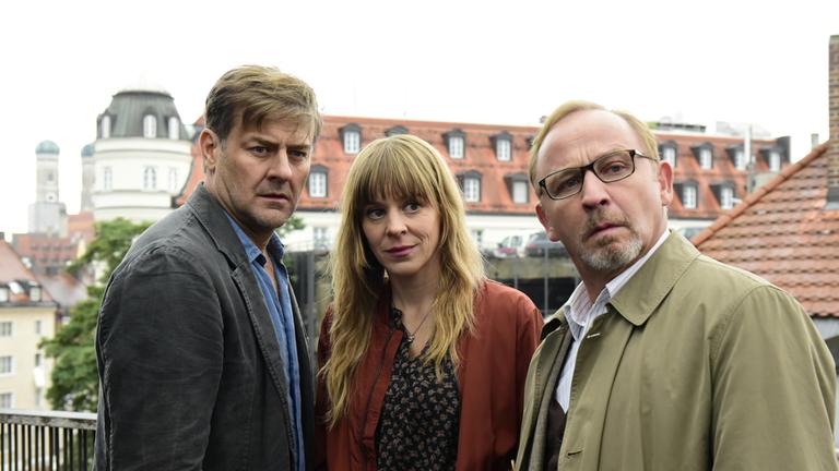 "München Mord – Auf der Straße, nachts, allein": Die drei Kommissare Harald Neuhauser (Marcus Mittermeier, l.), Angelika Flierl (Bernadette Heerwagen, M.) und Ludwig Schaller (Alexander Held, r.) stehen zusammen auf einer Terrasse und blicken an der Kamera vorbei.