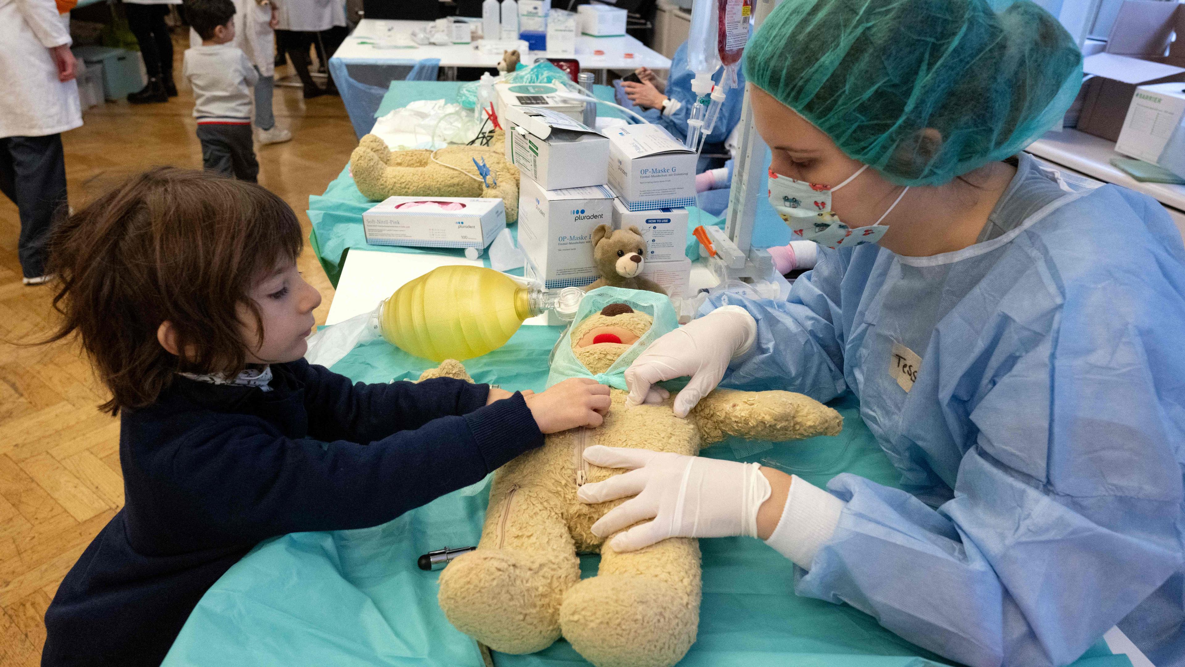 Eine Frau im Krankenhauskittel, Arzthandschuhen, Haarhaube und Mundschutz sitzt am Tisch einem kleinen Kind gegenüber. Auf dem Tisch liegt ein Teddybär. Auch er hat eine Patientenhaube an. Die Krankenhausschwester und das Kind schauen und fassen den Teddybär an.