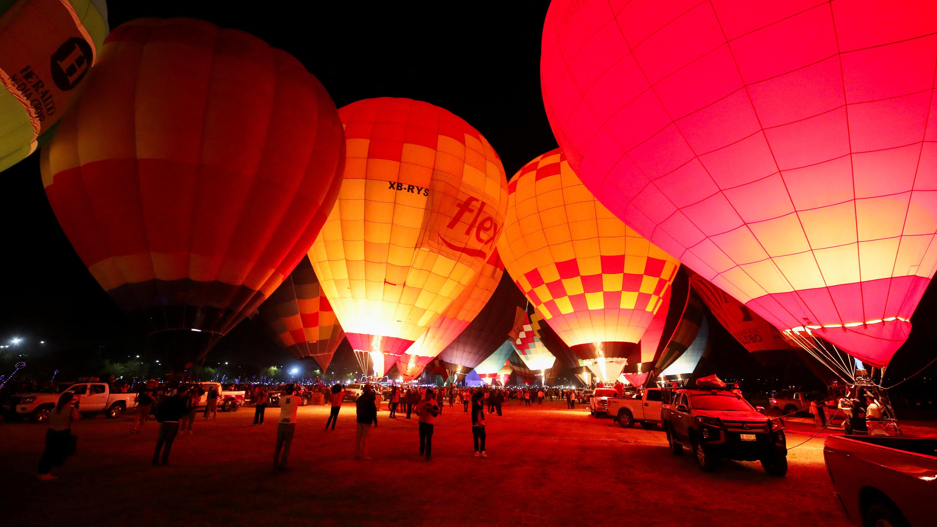 Rot leuchtende Heißluftballons am Boden bei Nacht, viele Menschen beobachten das Geschehen, ein paar Autos stehen daneben