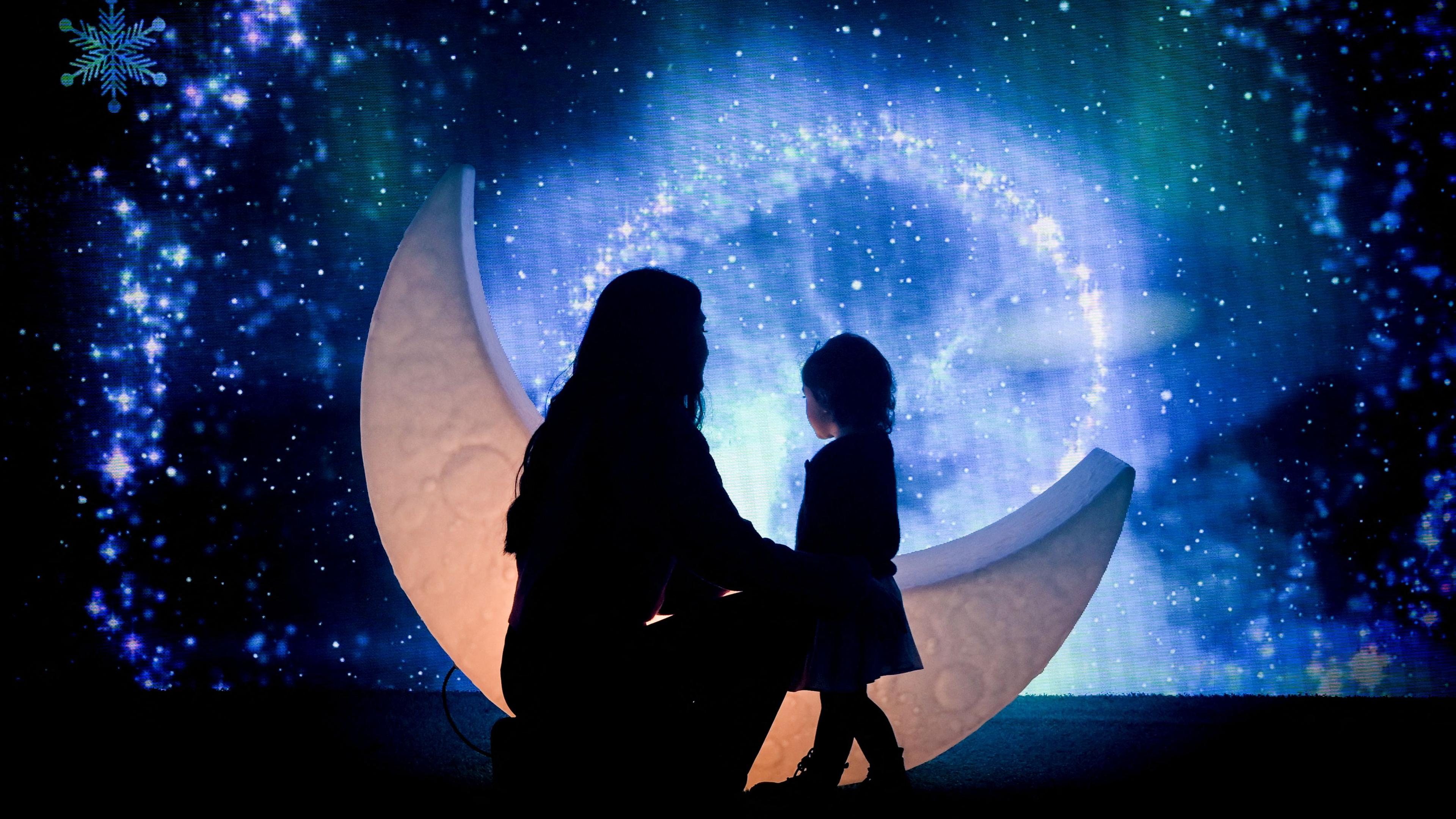 Die dunklen Umrisse von einer Frau und einem kleinen Mädchen sind vor einem sichelförmigen Mond vor einem mit großen Schneeflocken umsäumten glitzernden Sternenhimmel zu sehen. Der Hintergrund ist offensichtlich künstlich - als Attrappe gebastelt oder auf einem Computerbildschirm erzeugt. Die Silhpuette der Frau hat sich zum Mädchen hinuntergekniet, das ihr zugewandt sehr süß in Mantel und Stiefel gekleidet ist.