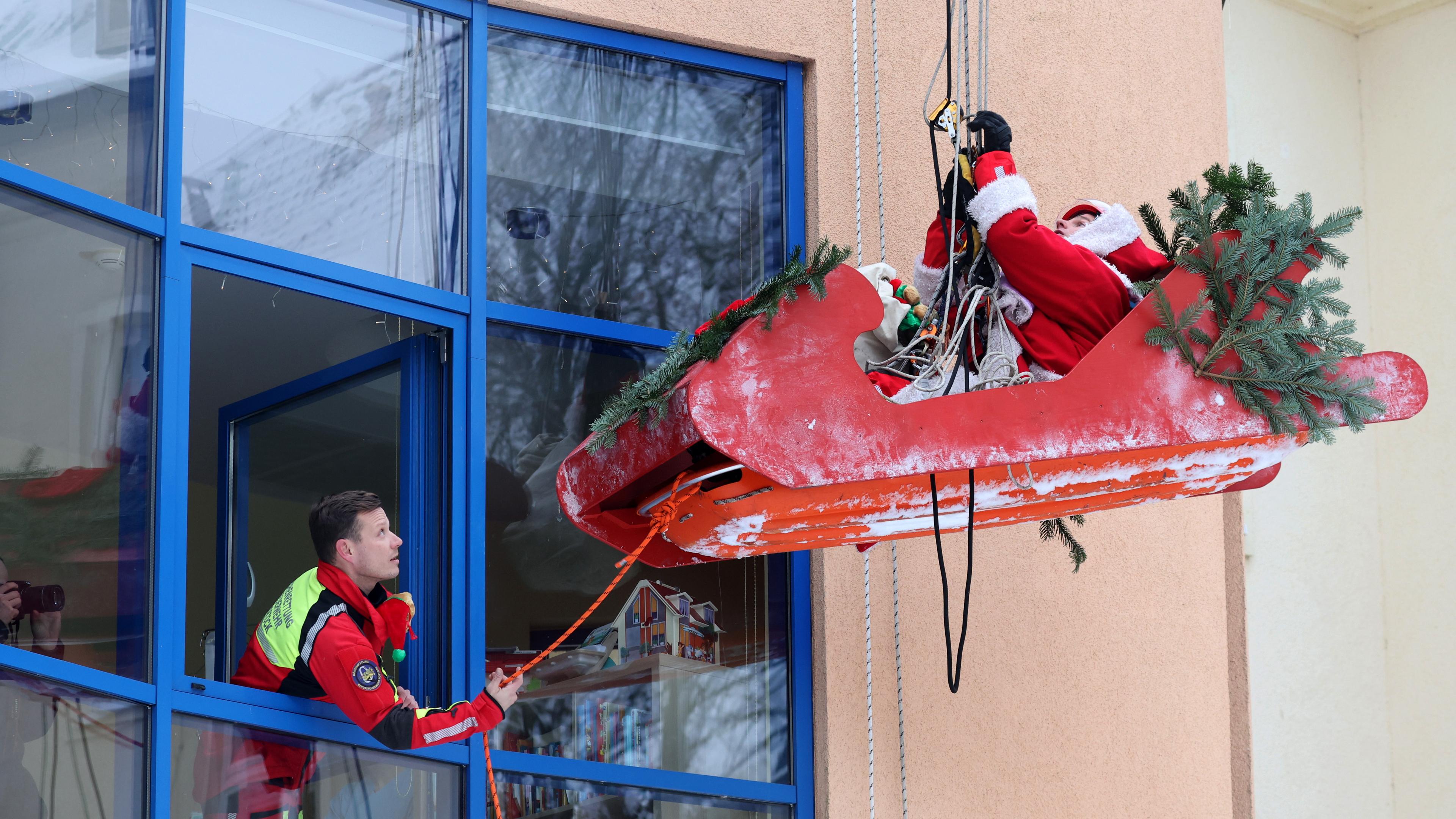 Wir sehen eine Hausfassade aus dessen geöffneten Fenstere sich ein Mann in Rettungskleidung beugt, und an einer Schnur zieht. Die Schnur führt zu einem Weihnachtsschlitten, indem ein Weihnachtsmann sitzt, der sich gerade samt schlitten abseilt.