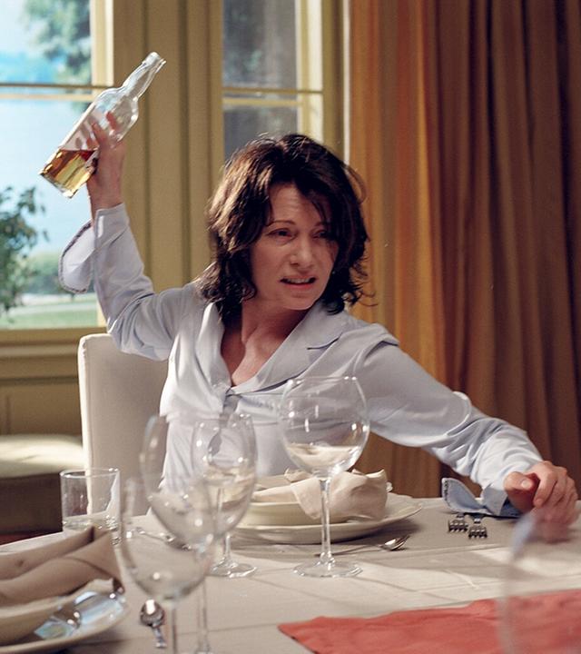 "Die Patriarchin": Nina Vandenberg (Iris Berben)sitzt am Tisch vor leeren Weingläsern, eine halbvolle Weinflasche in der erhobenen rechten Hand.  