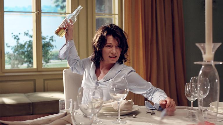 "Die Patriarchin": Nina Vandenberg (Iris Berben)sitzt am Tisch vor leeren Weingläsern, eine halbvolle Weinflasche in der erhobenen rechten Hand.  