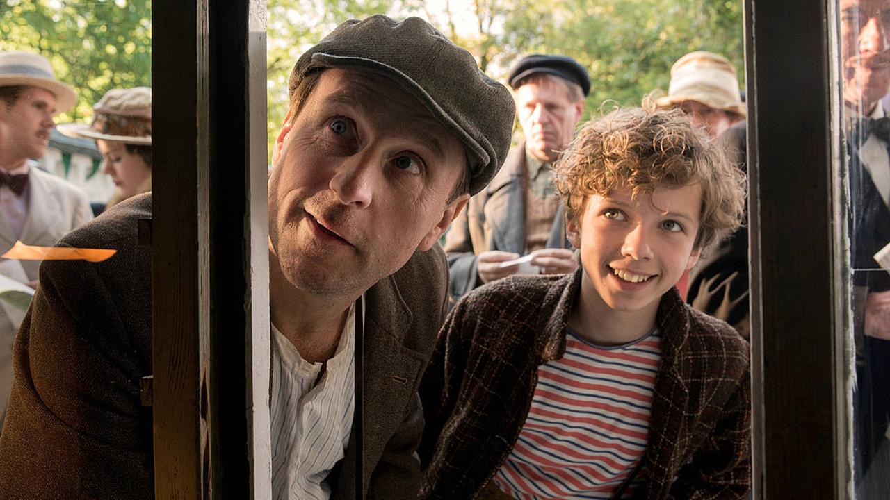 "Timm Thaler oder das verkaufte Lachen": Timm Thaler (Arved Friese) und Baron Lefuet (Justus von Dohnányi) schauen von außen durch ein Fenster. Im Hintergrund Passsanten.