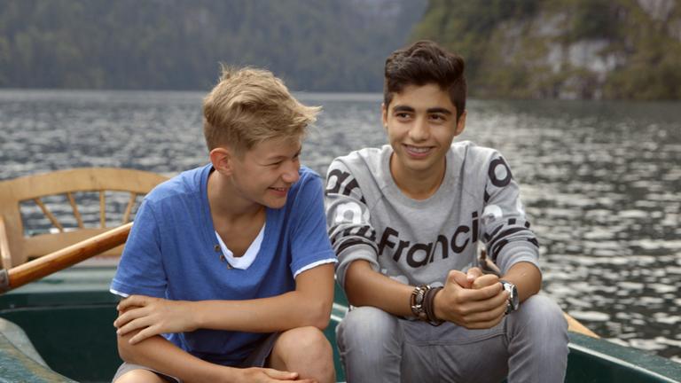Jeremia und Ibrahim - Brüder auf Zeit: Jeremia und Ibrahim (von links) sitzen nebeneinander in einem Ruderboot auf dem Wasser. Sie schauen in verschiedene Richtungen und lächeln.