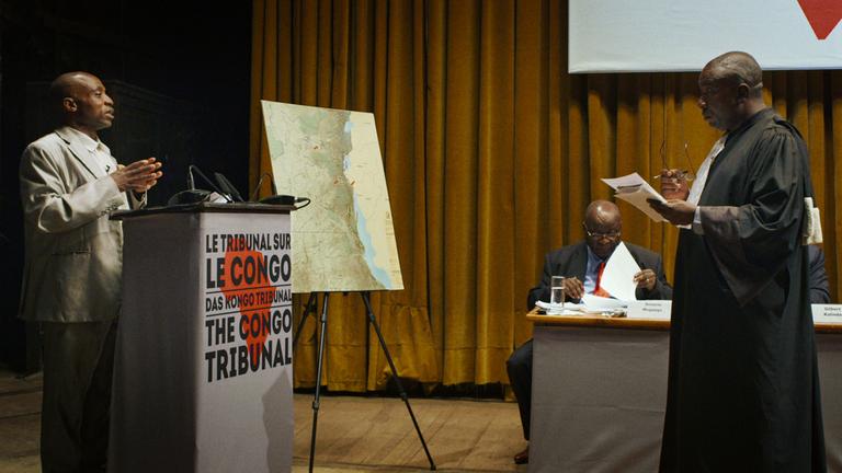 "Das Kongo-Tribunal": Links ein Mann hinter einem Stehpult mit der Aufschrift "Le Tribunal sur Kongo". Ihm gegenüber steht ein Mann in Richterrobe. Im Hintergrund vor einem gelben Vorhang links ein Flipchart mit einer Landkarte, rechts sitzt ein Mann an einem Tisch und studiert Papiere.