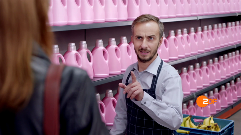 Spot für den Mediathek-Relaunch 2016: Jan Böhmermann als Supermarkt-Mitarbeiter vor einem Regal mit lauter gleichen rosa Waschmittel-Flaschen ohne Etiketten und einer Kiste Bananen. Im Vordergrund links eine Kundin.