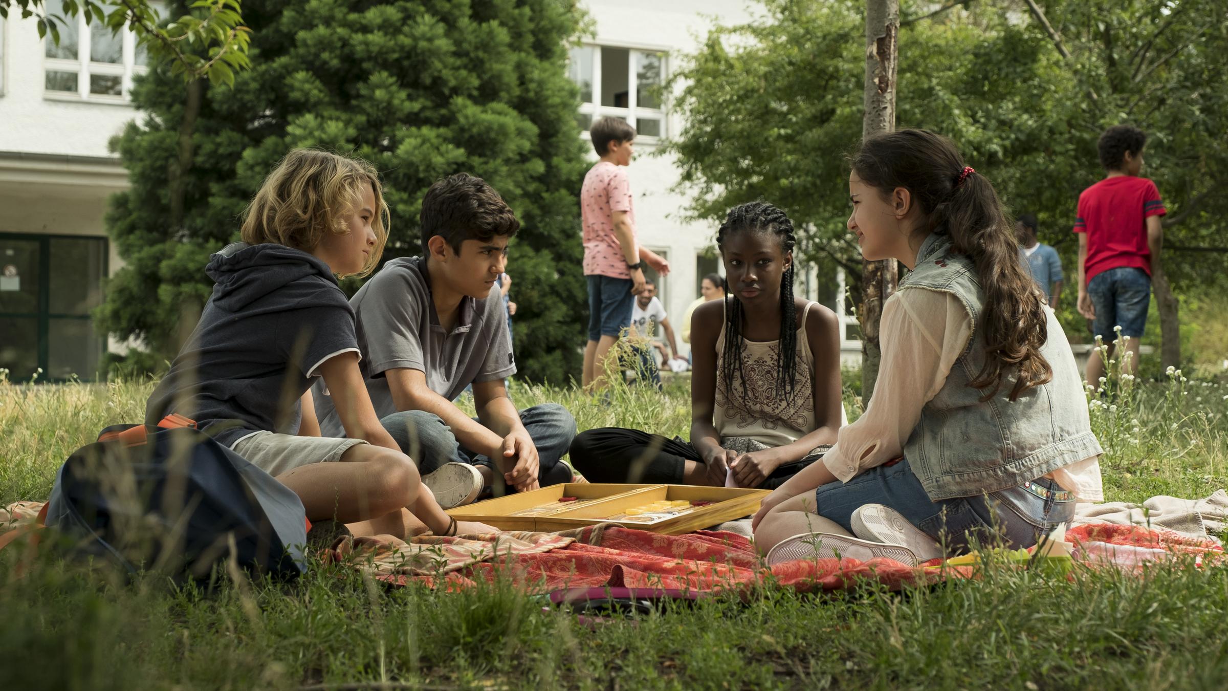 Dschermeni "Gegenüberstellungen": Moritz, Yassir, Aminata und Rüyet sitzen zusammen auf der Wiese vor einem Flüchtlingsheim.