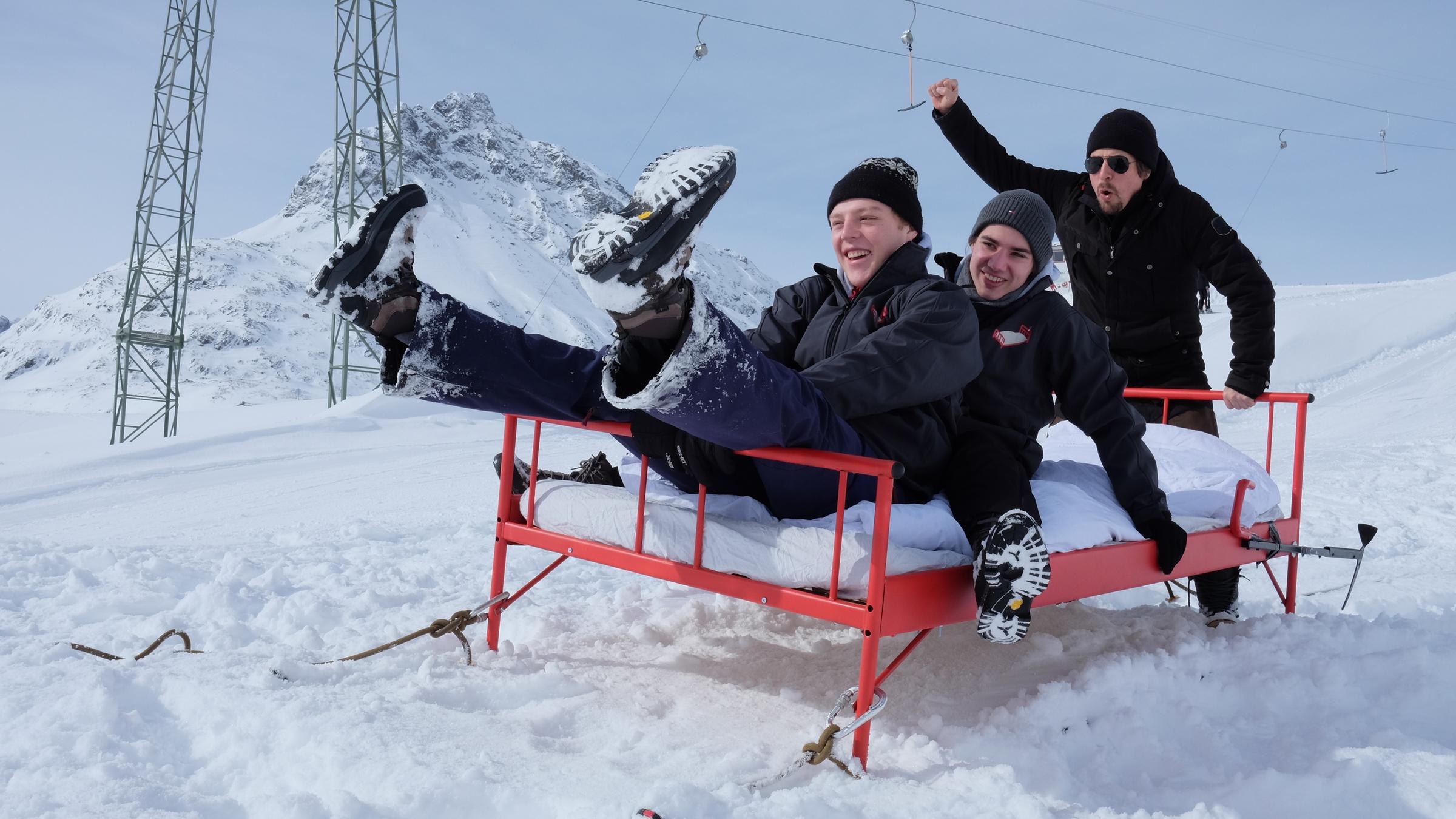 "Draußen schlafen - der Bettkampf", Kenneth, Leopold und Moderator Tommy Scheel auf einem roten Bett im Schnee vor verschneiter Bergkulisse und Schlepplift.