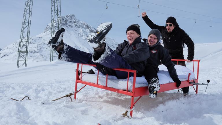"Draußen schlafen - der Bettkampf", Kenneth, Leopold und Moderator Tommy Scheel auf einem roten Bett im Schnee vor verschneiter Bergkulisse und Schlepplift.