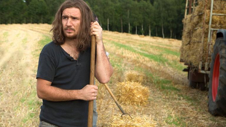 "Gutland": Jens (Frederick Lau) steht, gestützt auf eine Heugabel auf einem Feld neben einem Traktor mit Heuanhänger und schaut skeptisch.