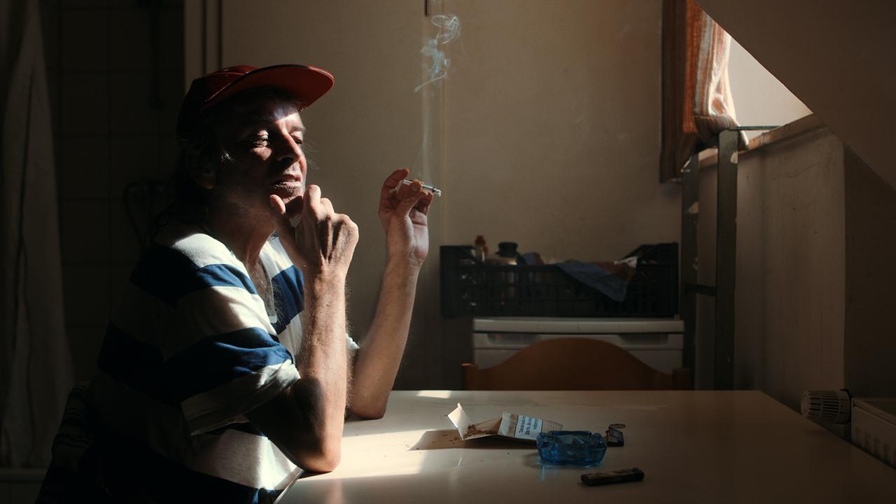 "SUPER FRIEDE LIEBE LOVE": In einem Obdachlosenheim sitzt ein Mann mit den Ellenbogen auf einen Tisch gestützt. In der linken Hand eine Zigarette. Auf dem Tisch Zigarettenpackung, Aschenbecher und Feuerzeuug. Durch ein kleines Fenster fällt etwas Sonnne ins Zimmer.