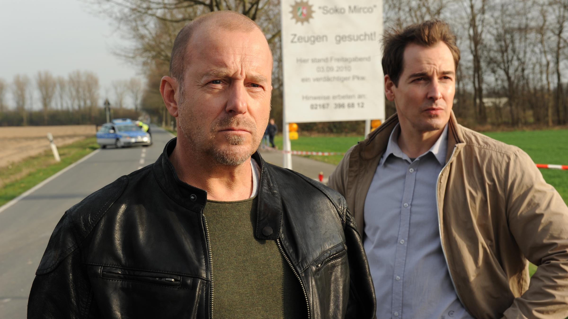 "Ein Kind wird gesucht": Die Ermittler Ingo Thiel (Heino Ferch) und Mario Eckartz stehen auf einer abgesperrrten Landstraße. Im Hintergrund rechts ein Fahndungsplakat mit Zeugenaufruf, links ein Polizeiwagen.