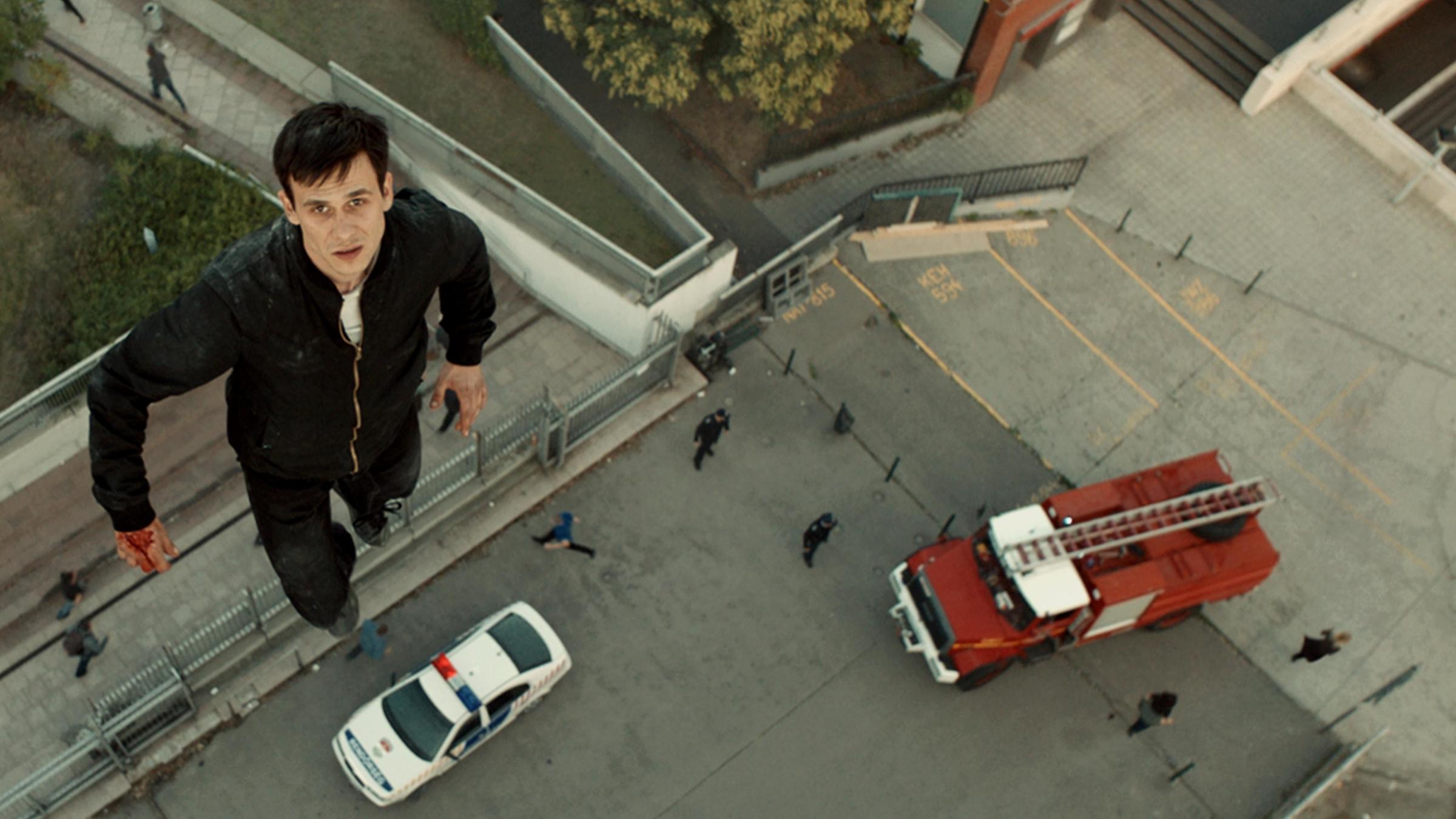 "Jupiter's Moon" (ARTE): Der junge Flüchtling Aryan (Zsombor Jéger) fliegen. Er schwebt aufrecht und schaut dabei in die Kamera. Unter ihm stehen auf einem betonierten Parkplatz ein Polizei- und ein Feuerwehrwagen.