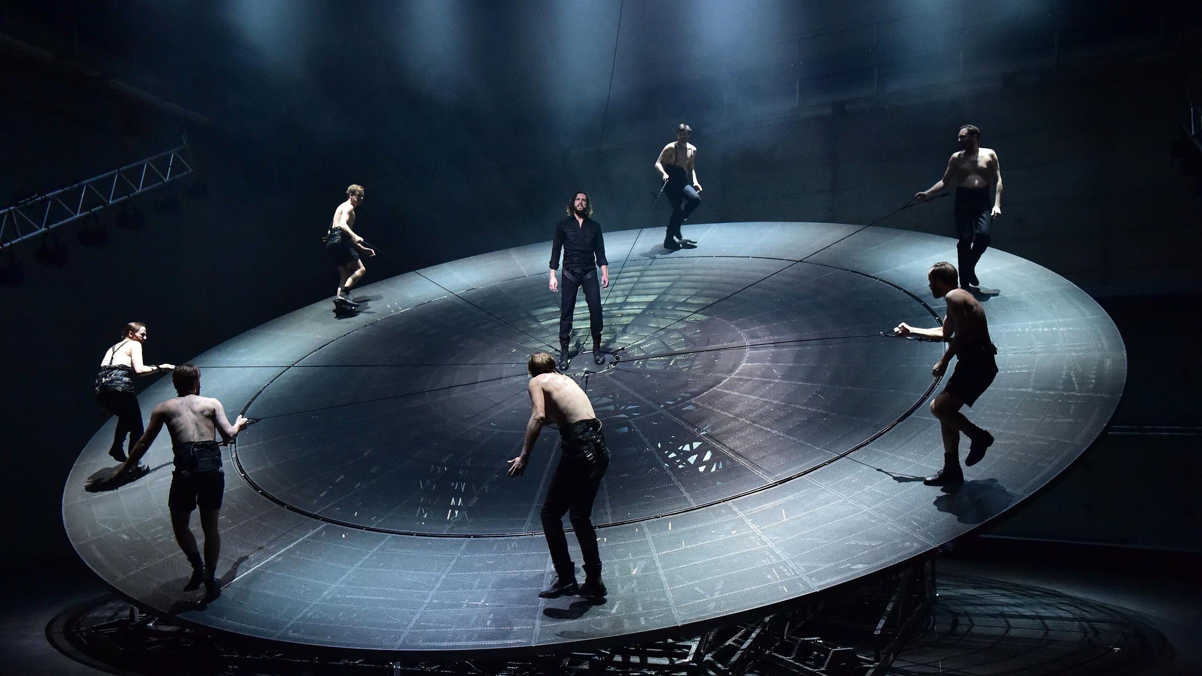 "Woyzeck" - Ein schwarz gekleideter Mann (Nicola Mastroberardino) steht inmitten einer riesigen metallenen, leicht nach vorne gekippten Scheibe. Um ihn herum sechs weitere Personen mit nacktem Oberkörper am Rand der Scheibe stehend und durch Seile gesichert.