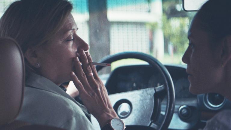 Chela (Ana Brun) sitzt rauchend am Steuer eines Autos und schaut dabei Angy (Ana Ivanova) auf dem Beifahrersitz an.