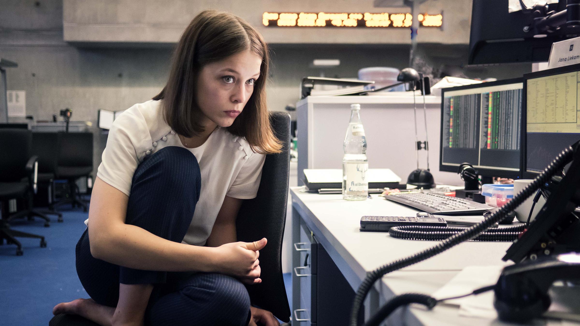 "Bad Banks": Jana Liekam (Paula Beer) sitzt mit angezogenen Beinen zusammengekauert auf dem Stuhl an ihrem Arbeitsplatz und schaut sorgenvoll