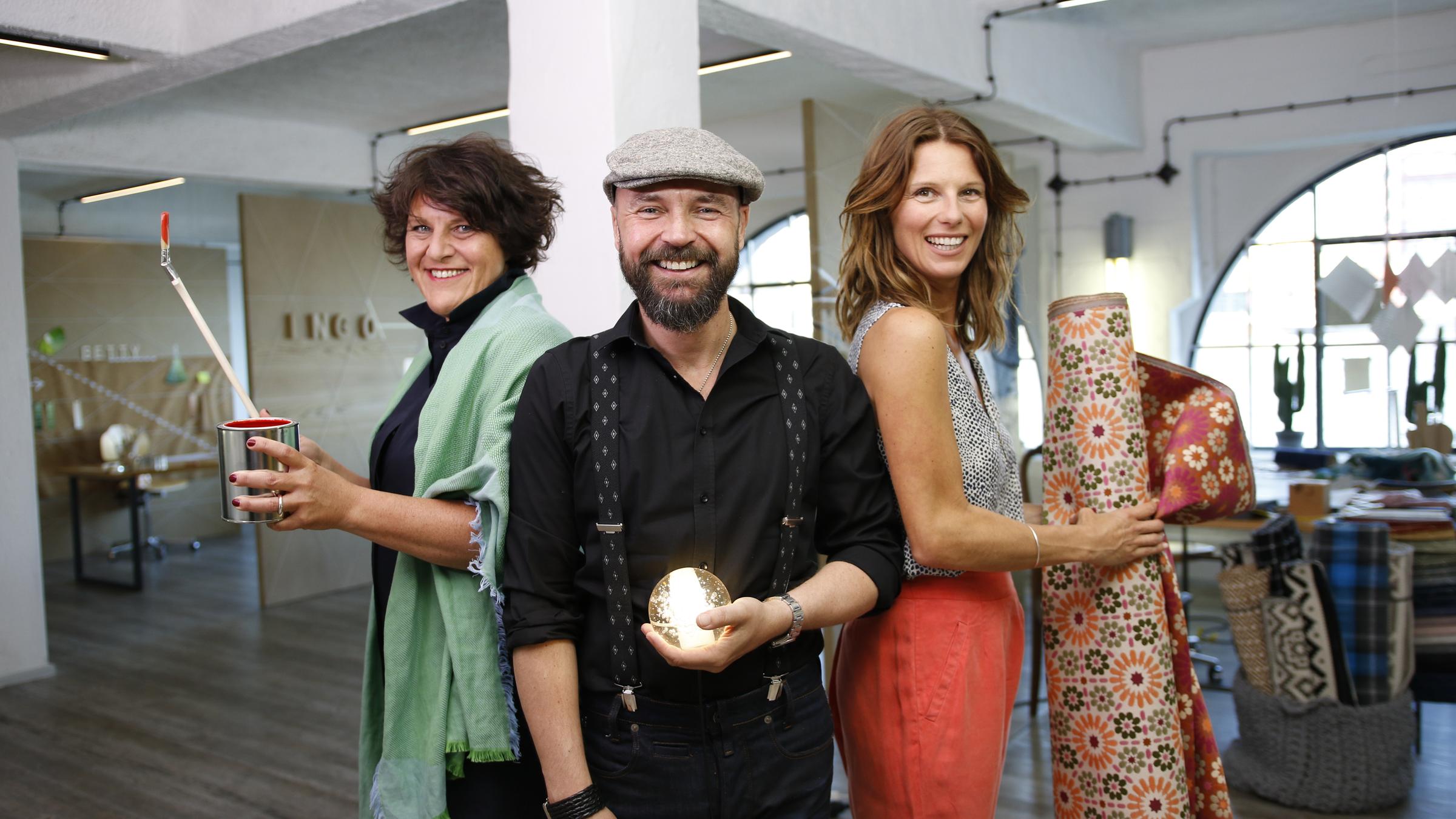 "3 Pläne für mein schönstes Zimmer": Betty, Ingo und Sasha stehen in einem Loft Rücken an Rücken im Halbkreis. Jede Person hält einen für sie typischen Gegenstand in der Hand (Farbtopf, Lampe, Stoffballen).