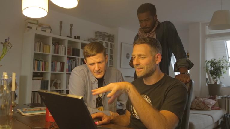 "Blacktape": Falk Schacht, Marcus Staiger und Sékou Neblett sitzen an einem Tisch vor einem aufgeklappten PC.