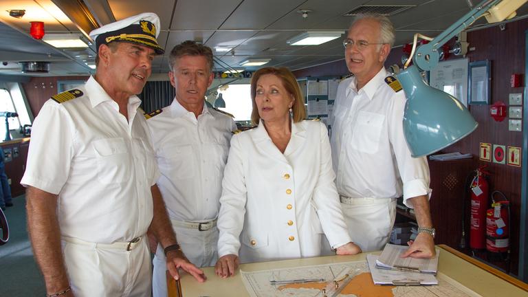 "Das Traumschiff - Palau": Kapitän Burger (Sascha Hehn), Dr. Sander (Nick Wilder), Beatrice (Heide Keller) und Oskar Schifferle (Harald Schmidt) stehen an einem Tisch, auf dem eine Seekarte ausgebreitet liegt, und reden miteinander.