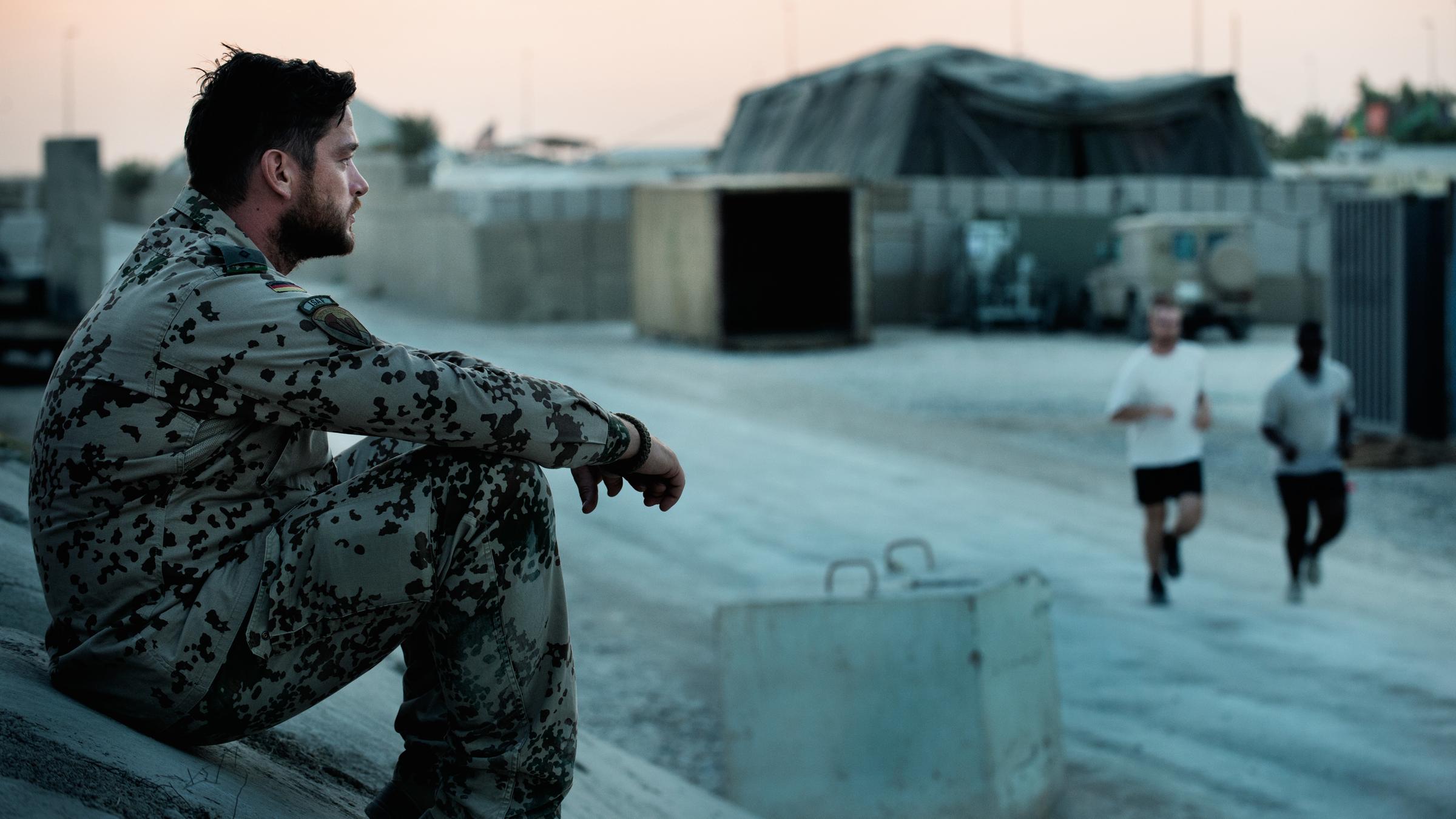 Zwischen Welten": Jesper (Ronald Zehrfeld) sitzt in Bundeswehrkleidung auf einem Hügel vor einem Gelände, auf dem ein großes Zelt steht. Er blickt nachdenklich geradeaus. Zwei Jogger laufen an ihm vorbei.