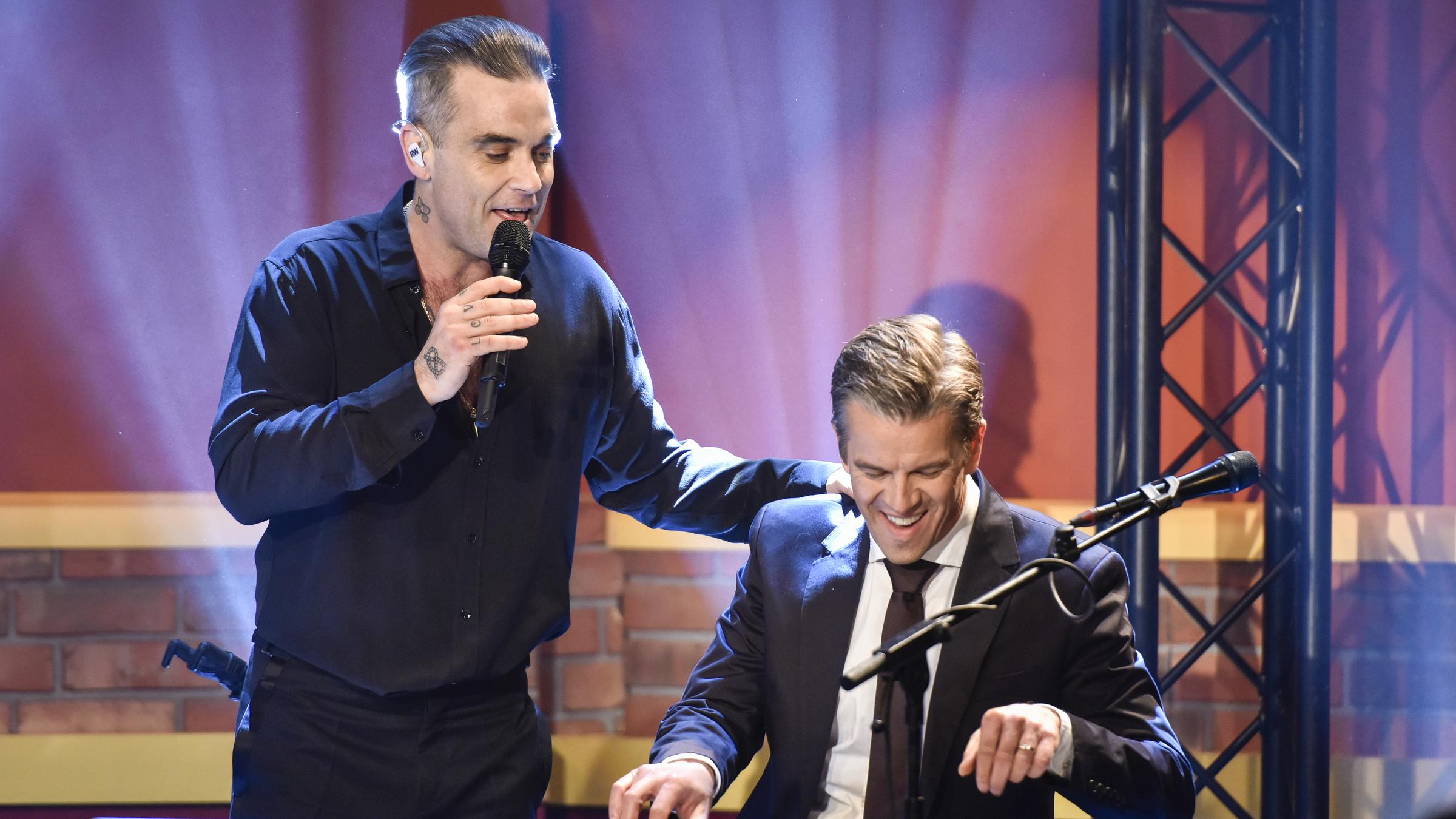"Markus Lanz": Robbie Williams hält ein Mikrofon in der Hand und singt, Markus Lanz begleitet ihn am Klavier.