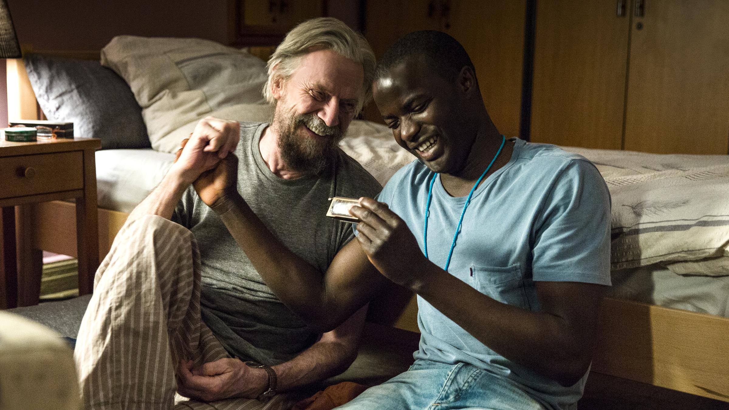 "Der Andere - Eine Familiengeschichte": Flüchtling Nama (Nama Traore) sitzt neben Willi (Jesper Christensen) im Seniorenheim. Sie unterhalten sich lachend.