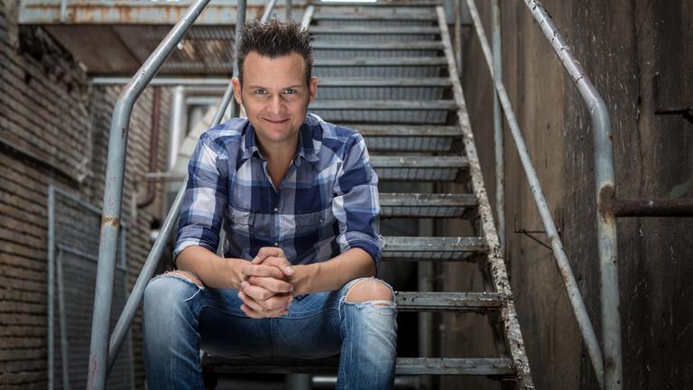 "3satfestival - Die Jubiläumsgala": Tobias Mann im karierten Hemd sitzt auf einer Treppe.
